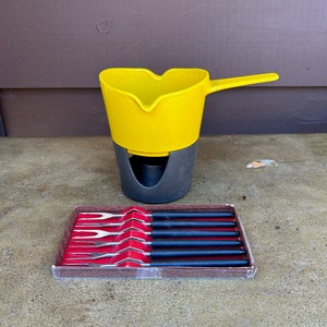 Könnte beinhalten: Ein gelber Fonduetopf mit einem schwarzen Ständer und einem Satz von sechs Fonduegabeln in einer roten und braunen Schachtel.