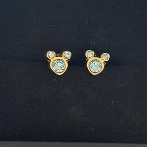 May include: A pair of gold-coloured stud earrings shaped like Mickey Mouse's head. Each earring has a light blue crystal for the face and two smaller crystals for the ears. The earrings are set against a dark background.