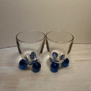 Può includere: Due bicchieri di vetro trasparente con base arrotondata, ciascuno sostenuto da tre sfere di vetro blu cobalto. I bicchieri hanno un design semplice ed elegante, adatto per servire bevande. Lo sfondo è di colore neutro.