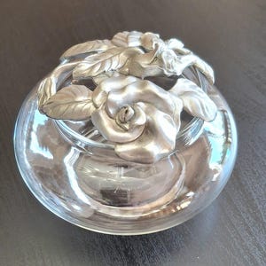 May include: A clear glass bowl featuring a decorative silver-toned metal lid. The lid is shaped like a cluster of sculpted roses and leaves, resting securely on the rim of the circular glass vessel. The metal has a matte, brushed finish.