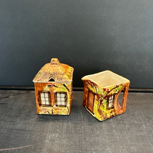 Puede incluir: Un juego vintage de azúcar y crema de cerámica con forma de cabaña. La jarra de crema tiene un asa y el azucarero tiene una tapa. Ambas piezas están decoradas con un diseño floral marrón, amarillo y verde.