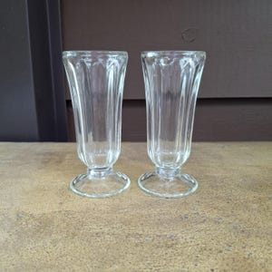 Puede incluir: Dos vasos de refresco transparentes de estilo vintage. Cada vaso tiene un diseño acanalado y una base redondeada. Los vasos son de vidrio transparente y se exhiben sobre una superficie marrón claro.