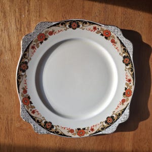 May include: White ceramic plate with a decorative floral border in black, orange, and gold. The plate has a scalloped edge and a simple, elegant design, suitable for serving food or as a decorative piece.