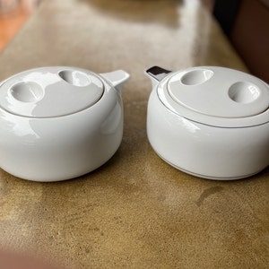 May include: Two white ceramic food storage containers with lids. The lids have two small circular openings. The containers have a silver trim around the top.
