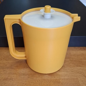 Può includere: Una brocca di plastica gialla con manico e coperchio bianco. La brocca ha un corpo arrotondato e un beccuccio. Il coperchio ha un piccolo pomello giallo. La brocca è su una superficie di legno.
