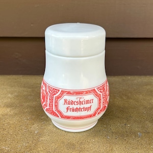 May include: A white ceramic jar with a lid, featuring a red decorative label with the text "Rüdesheimer Früchtetopf." The jar has a rounded shape and a slightly flared base. The label has an ornate design.