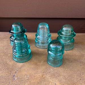 Peut inclure: Ensemble de cinq isolateurs en verre bleu-vert antiques. Chaque isolateur a un sommet arrondi et un corps conique strié. Le verre est légèrement transparent. Les isolateurs sont disposés sur une surface texturée. Composants électriques vintage.