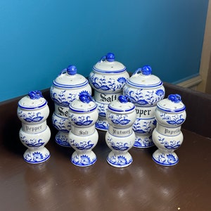 Puede incluir: Conjunto de ocho tarros de especias de cerámica azul y blanca con tapas. Los tarros están decorados con un patrón floral y tienen las palabras "Sal", "Pimienta", "Pimentón", "Nuez moscada" y "Sal marina" escritas en ellos.