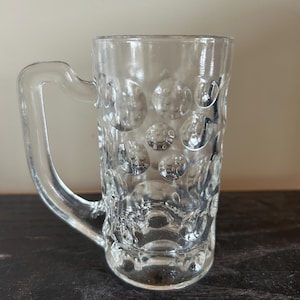 May include: A clear glass beer mug with a textured, bubble-like design. The mug has a handle and a rounded base.