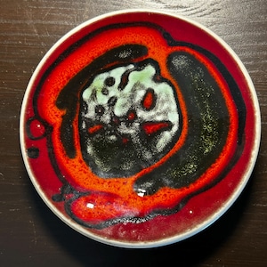 May include: A round ceramic plate with a white rim. The plate features a vibrant design with a red outer ring, a black inner ring, and a central area with green and white patterns. The plate is likely for decorative or serving purposes.