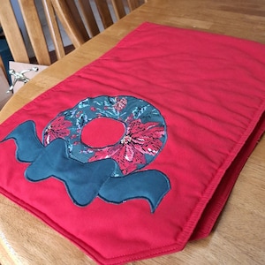 May include: A red table runner with a Christmas wreath design. The wreath is teal with red poinsettias and a teal bow. The runner is rectangular and made of fabric.
