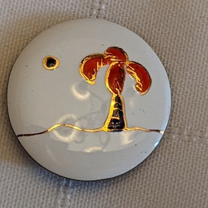 May include: A white ceramic button featuring a gold palm tree design. The palm tree has red leaves and a black trunk. There is a small gold circle in the top left corner of the button.