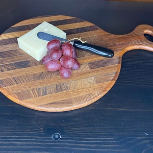 Puede incluir: Una tabla de cortar redonda de madera con un asa, con un patrón a rayas. La tabla está cubierta con un bloque de queso, un pequeño cuchillo y un racimo de uvas rojas.