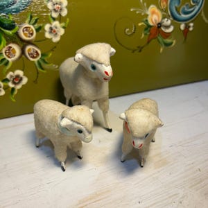 Puede incluir: Tres pequeñas figuras de ovejas de estilo vintage. Las ovejas son blancas con pezuñas negras, ojos azules y detalles rojos en la nariz. Están dispuestas sobre una superficie blanca con un fondo verde y floral pintado.