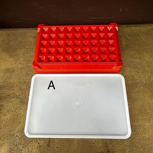 Op de afbeelding: Een rechthoekige rode plastic ijsblokjesvorm met een raster van vierkante vakjes en een apart, ondoorzichtig wit rechthoekig deksel. Het deksel is voorzien van de letter A in het zwart aan de linkerkant en een kleine ronde inkeping in het midden.