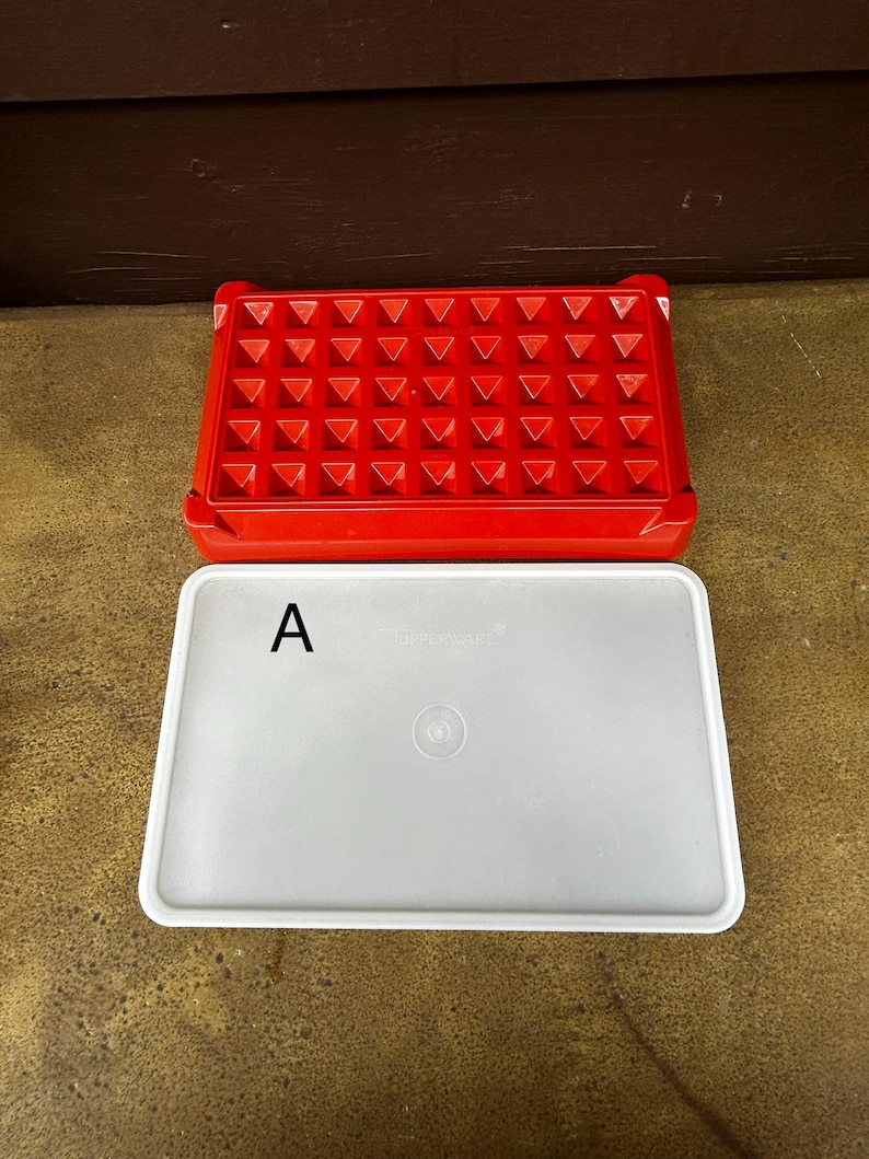 Op de afbeelding: Een rechthoekige rode plastic ijsblokjesvorm met een raster van vierkante vakjes en een apart, ondoorzichtig wit rechthoekig deksel. Het deksel is voorzien van de letter A in het zwart aan de linkerkant en een kleine ronde inkeping in het midden.