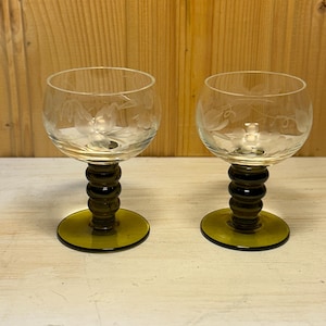 Puede incluir: Dos copas de vino transparentes con diseños florales grabados. Los tallos son de color verde oscuro con un diseño bulboso, y las bases son de color verde a juego. Las copas se exhiben sobre una superficie clara.