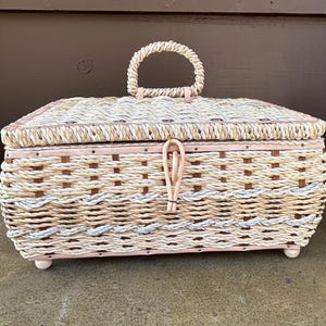 May include: A woven wicker storage box with a light pink painted finish and a rope handle. The box has a woven pattern with a light brown and white color scheme.