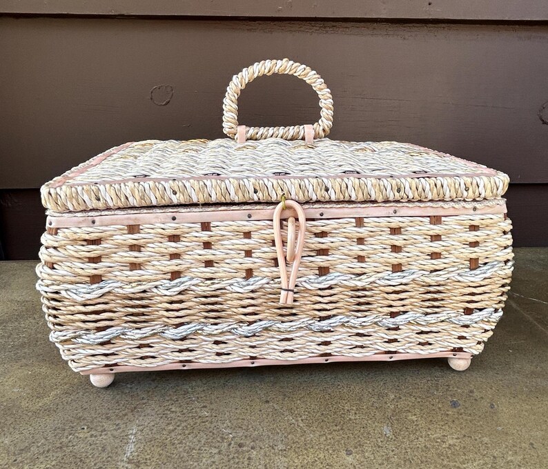 May include: A woven wicker storage box with a light pink painted finish and a rope handle. The box has a woven pattern with a light brown and white color scheme.