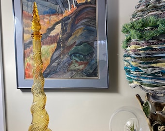 May include: A tall, amber-colored glass bottle with a spiral design. The bottle has a bulbous base and a long, twisted neck. The bottle is displayed on a dark surface, with a framed watercolor painting in the background.