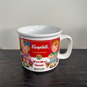 May include: White ceramic mug with a Campbell's Condensed Soup design. The mug has a red band with the Campbell's logo and illustrations of children. The text "M'm! M'm! Good! Soup" is also displayed on the mug.