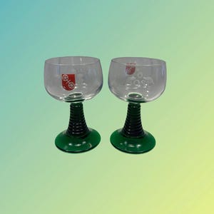 Könnte beinhalten: Zwei klare Weingläser aus Glas mit grünen Stielen und Füßen. Die Gläser haben ein rotes Logo mit einem Radmotiv auf der Vorderseite.
