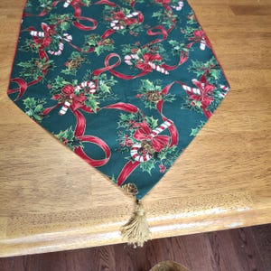 Puede incluir: Un camino de mesa navideño verde con un patrón festivo de lazos rojos, bastones de caramelo, acebo y piñas. El camino de mesa tiene una borla dorada en el extremo, añadiendo un toque de elegancia.