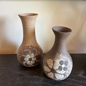 May include: Two ceramic vases with a floral design. The vases are brown and tan with a bulbous base and a narrow neck. The floral design is in black and white.