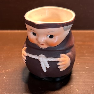 Puede incluir: Figura de cerámica de un monje, con una túnica marrón, un cinturón de cuerda blanco y una cara sonriente con mejillas rosadas. El pequeño recipiente abierto es probablemente un artículo decorativo.