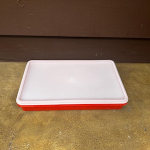May include: A rectangular storage container with a bright red base and a translucent white lid. The lid features a centred circular design and a raised rim, while the base has straight sides and a flat bottom, creating a simple food storage unit.