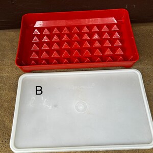 Op de afbeelding: Een rechthoekig rood plastic dienblad met een raster van kleine driehoekige inkepingen en een apart doorschijnend wit plastic deksel. Het deksel is voorzien van de letter B en de merknaam Tupperware in reli&euml;f.