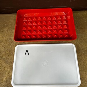 Op de afbeelding: Een rechthoekig rood plastic dienblad met een raster van verhoogde driehoekige inkepingen op de bodem, gecombineerd met een apart, doorschijnend wit rechthoekig deksel. Het deksel is gemarkeerd met de letter A en de merknaam Tupperware.