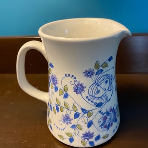 Może przedstawiać: Biały dzbanek ceramiczny z niebieskim i zielonym wzorem kwiatowym przedstawiającym stylizowane postaci. Dzbanek ma uchwyt i dzióbek.