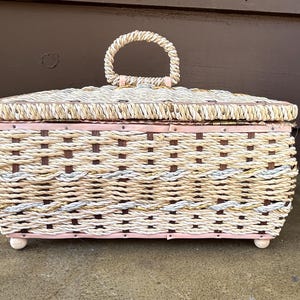 May include: A woven wicker storage box with a pink painted base and a copper colored metal handle. The box has a woven pattern with brown and white rope.