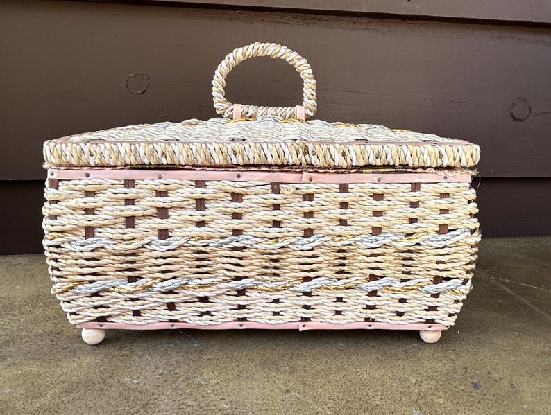 May include: A woven wicker storage box with a pink painted base and a copper colored metal handle. The box has a woven pattern with brown and white rope.