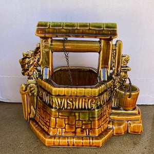 Può includere: Un vaso da fiori in ceramica a forma di pozzo con una smaltatura marrone e verde. Il pozzo ha una catena e un secchio, e le parole "FOR VISITING" sono scritte sul davanti.