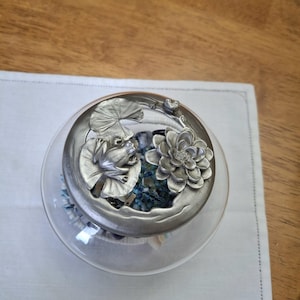 Puede incluir: Un frasco de vidrio redondo con una tapa decorativa de metal plateado que presenta una rana esculpida sentada sobre una hoja de nenúfar junto a una flor de loto en flor. El interior del frasco está lleno de pequeñas piedras decorativas de color azul oscuro y blanco.