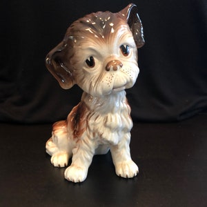 Puede incluir: Una figurita de cerámica de un perro marrón y blanco con una expresión triste. El perro está sentado con la cabeza inclinada hacia un lado y las patas metidas debajo del cuerpo.