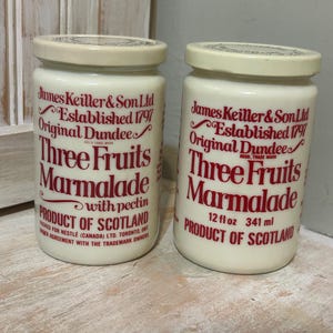 Puede incluir: Dos tarros de vidrio de mermelada de tres frutas, un producto de Escocia. Los tarros son blancos con letras rojas y un logotipo rojo. Los tarros son de 12 onzas líquidas o 341 mililitros.