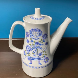 May include: White ceramic teapot with a lid, featuring a blue and purple floral design with a stylized figure. The teapot has a curved handle and a long spout. The design includes a border of blue dots around the base.