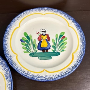 May include: White ceramic plate with a blue, yellow, and green design. The plate features a hand-painted illustration of a person in traditional clothing, surrounded by green foliage. The rim has a blue and white speckled pattern.