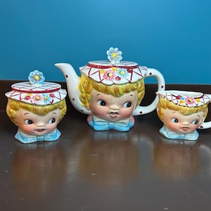 May include: A vintage ceramic tea set comprising a teapot, sugar bowl, and teacup, each decorated with a charming face design. The set is painted in yellow, white, and blue, with floral details and a red and white checkered pattern on the lids and rims.
