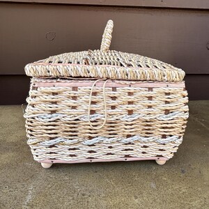 May include: A woven wicker basket with a pink painted base and a lid. The basket is made of natural colored and white rope.