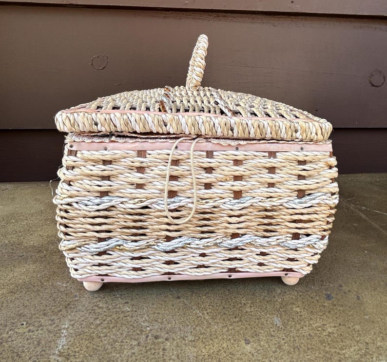 May include: A woven wicker basket with a pink painted base and a lid. The basket is made of natural colored and white rope.