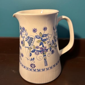 Może przedstawiać: Biały dzbanek ceramiczny z niebiesko-zielonym wzorem kwiatowym przedstawiającym stylizowaną kobietę w drzewie. Dzbanek ma uchwyt i żebrowaną podstawę.