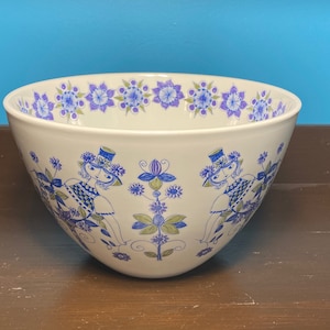 Pode incluir: Tigela de cerâmica branca com um padrão floral azul e roxo na borda. A tigela apresenta um design caprichoso de figuras com chapéus e motivos florais em azul e verde. A tigela tem uma forma ampla e aberta.