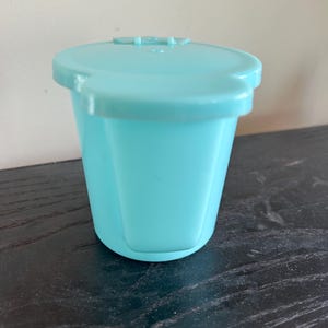 Puede incluir: Un recipiente de plástico azul claro con una tapa a juego. El recipiente tiene una forma ligeramente trapezoidal y una tapa con bisagras. La tapa tiene una pequeña pestaña para abrir. El recipiente está sobre una superficie de madera oscura.