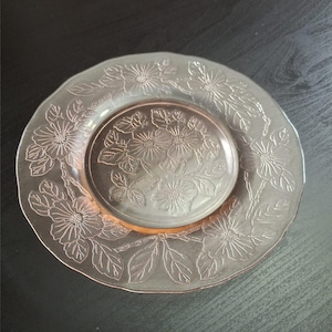 Puede incluir: Un plato de vidrio rosa con un diseño floral. El plato presenta flores y hojas en relieve alrededor del borde y el centro. El plato es redondo y tiene un borde ligeramente festoneado.