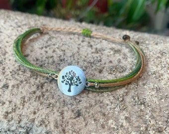 May include: A green and brown braided bracelet with a silver charm featuring a tree cutout design.
