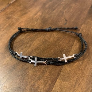 May include: A black braided cord bracelet with three silver cross charms.